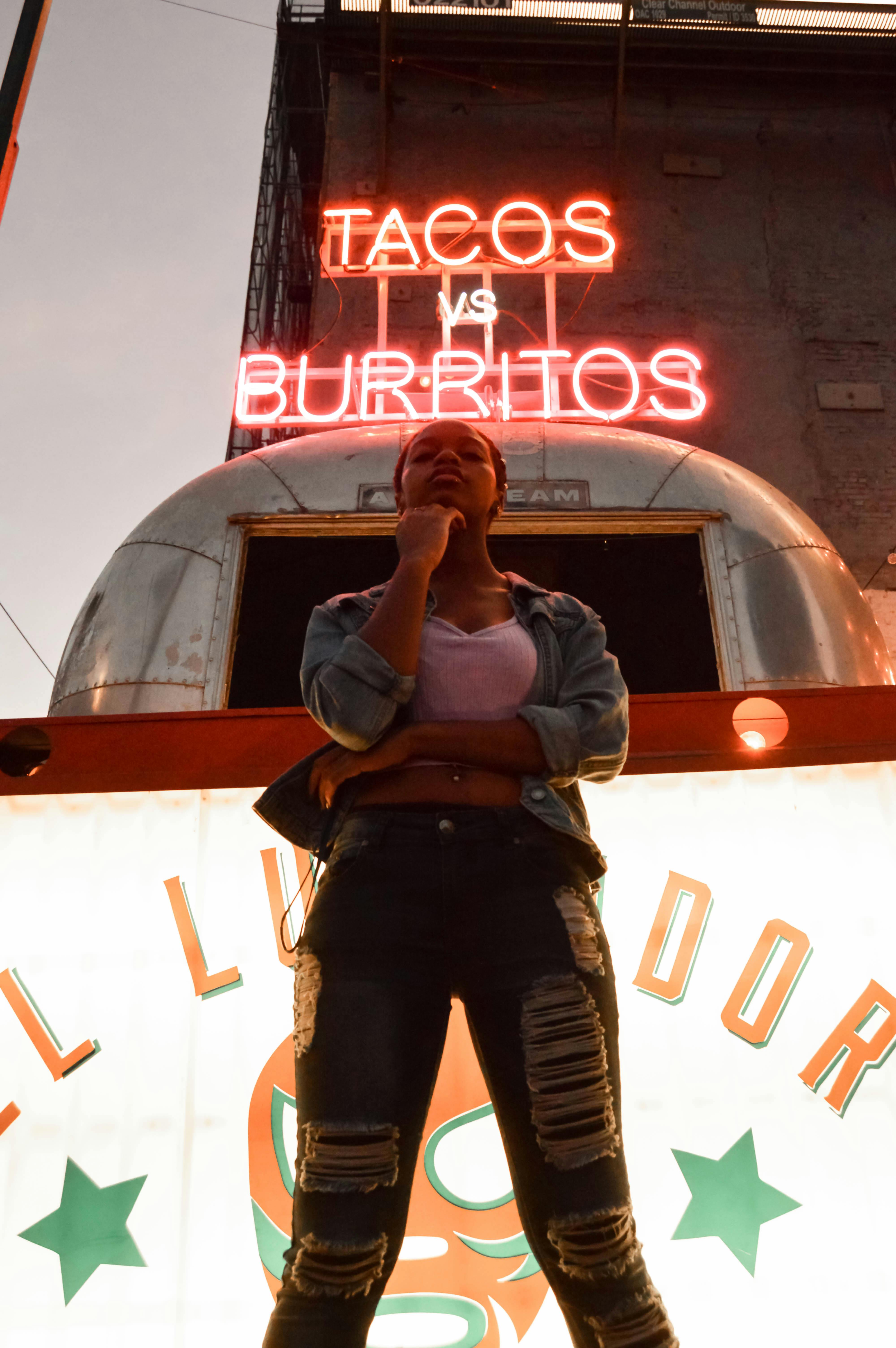 A woman stands thoughtfully under neon taco sign in urban setting.