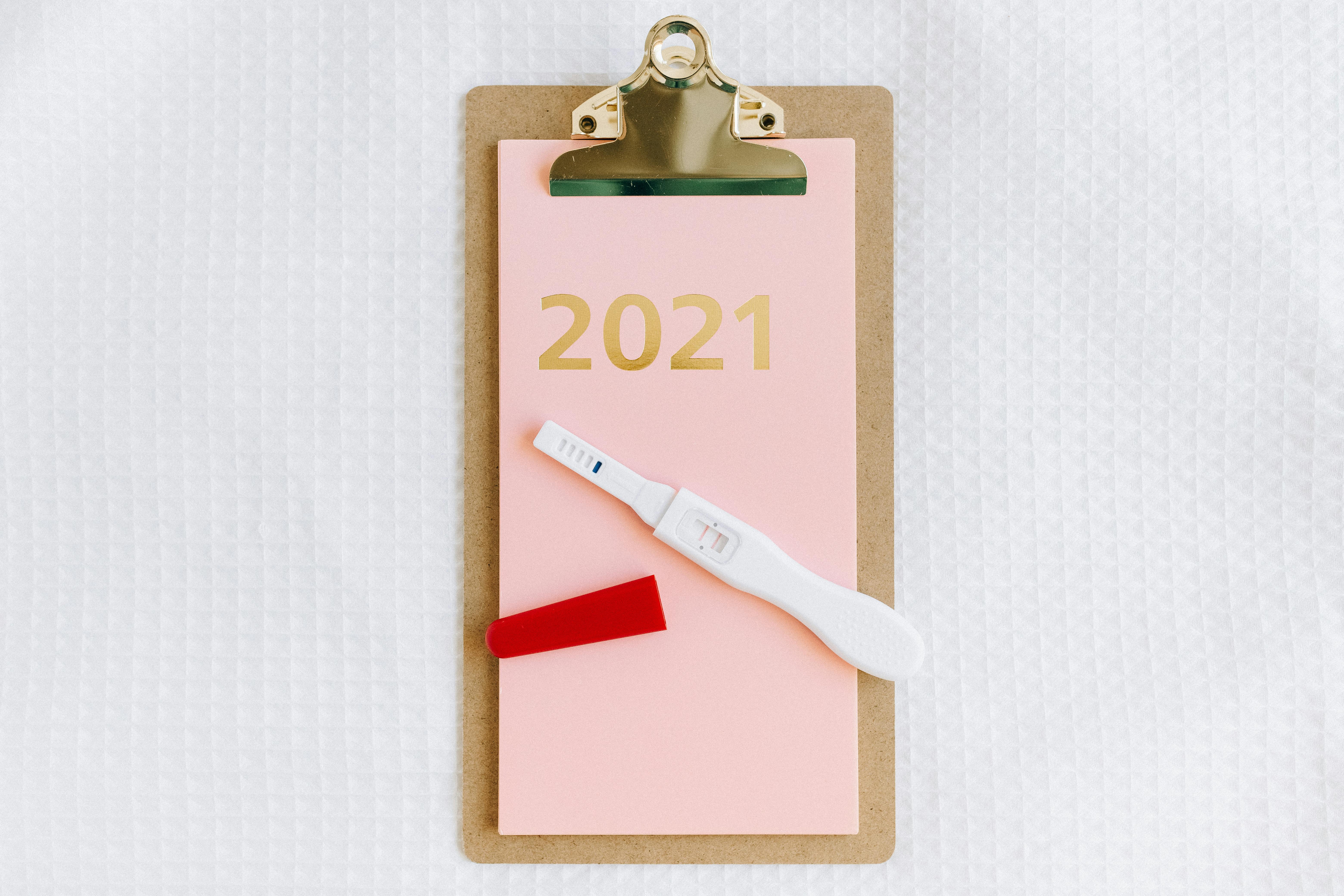 Flat lay image of a pregnancy test on a pink paper clipboard with 2021, offering copy space.