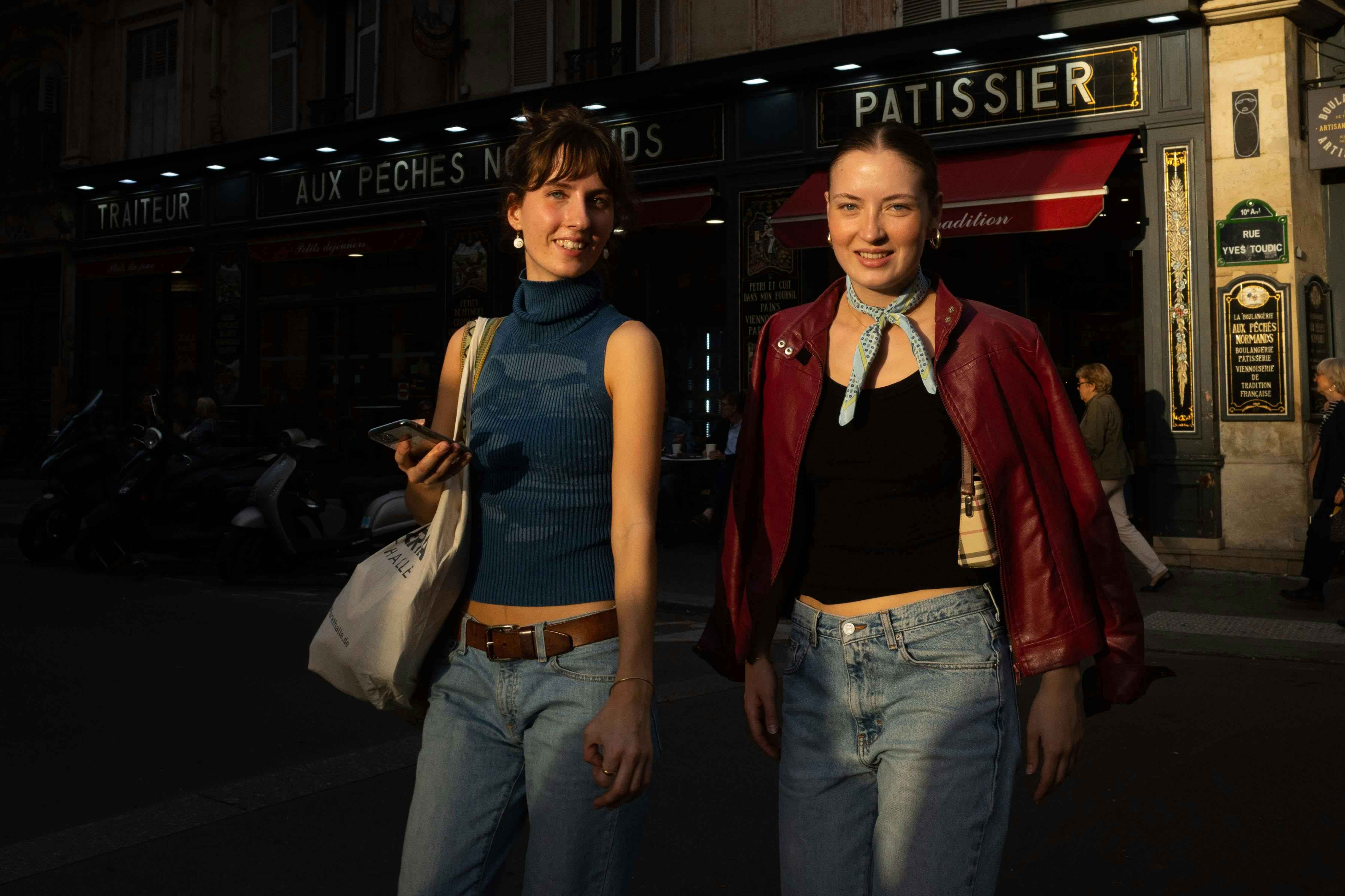 Two women walking on a city street near a Parisian patisserie during twilight, enjoying an evening stroll.