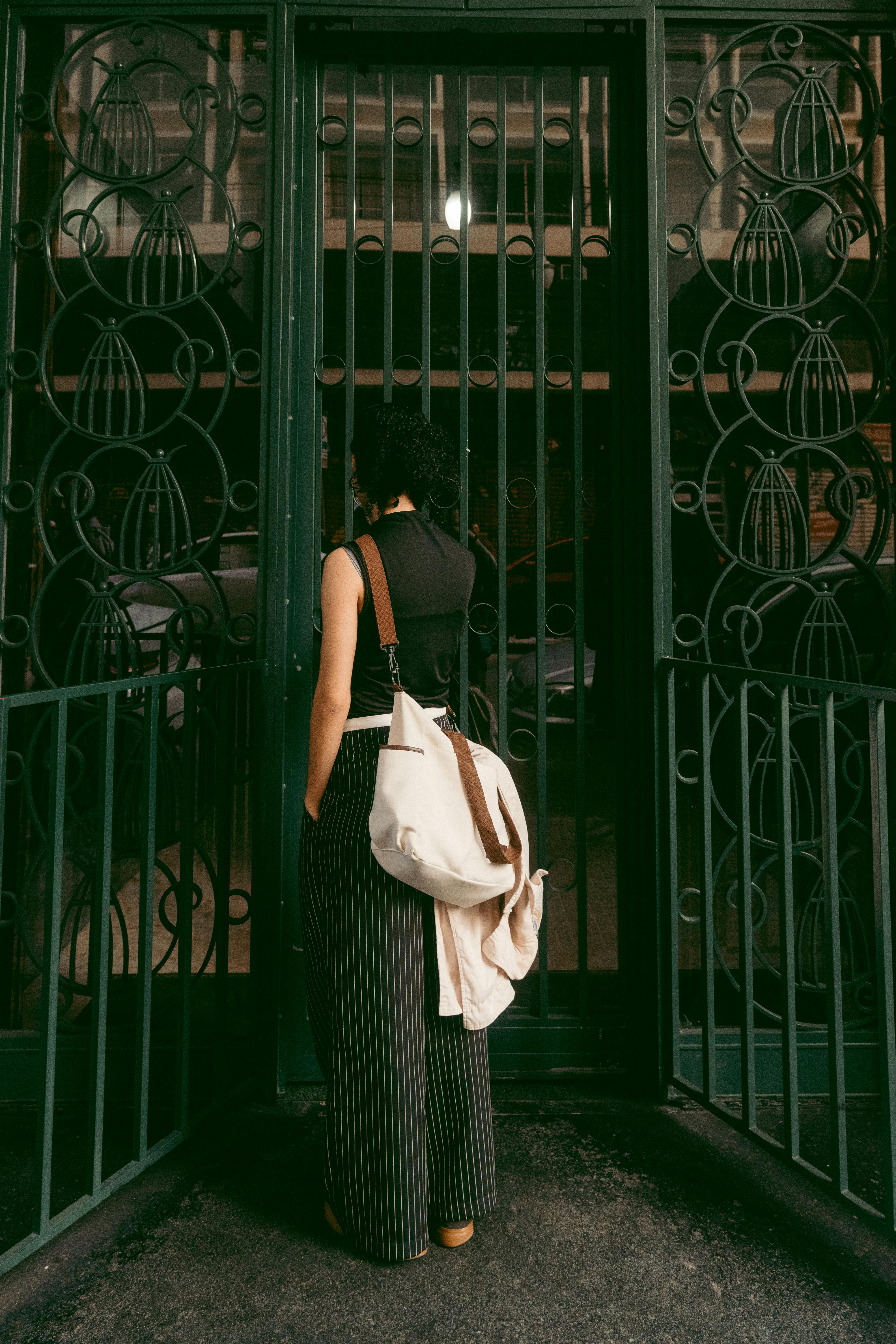 An individual stands at a decorative metal gate, viewed from behind, carrying a white bag.