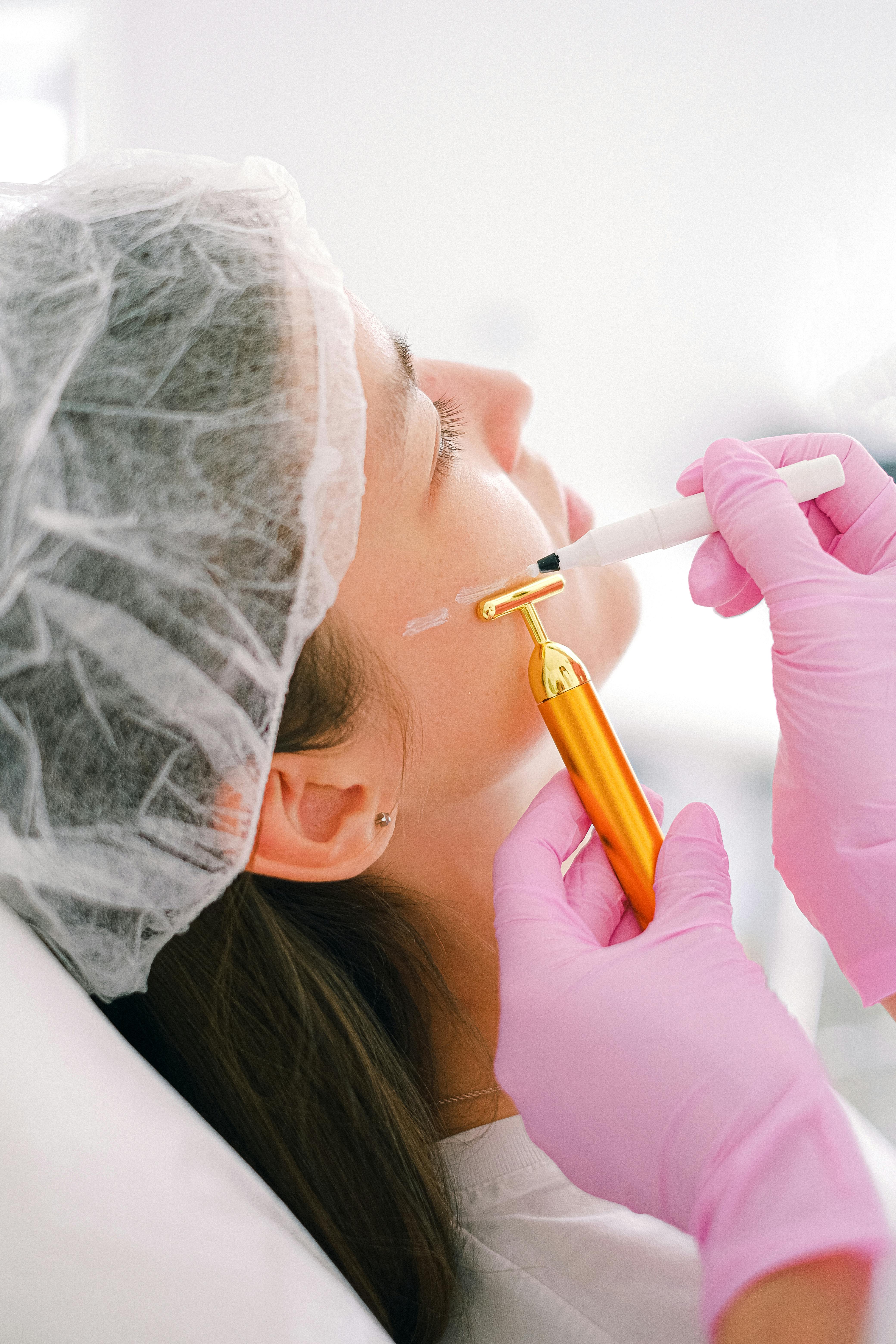 A cosmetologist performs a facial treatment using a golden massage roller and marker.