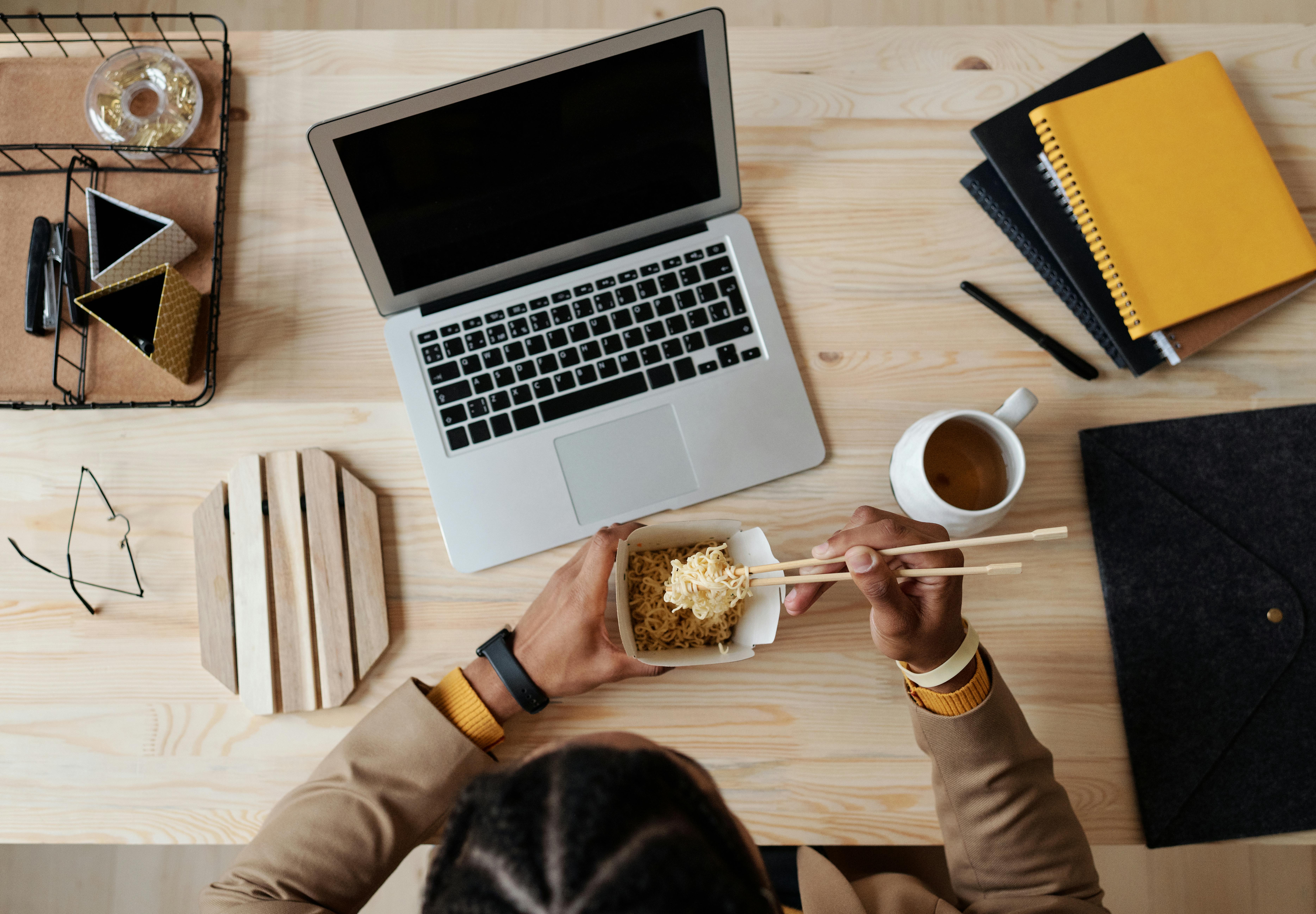 Top view of a person enjoying noodles while working on a laptop. Modern workspace setup.
