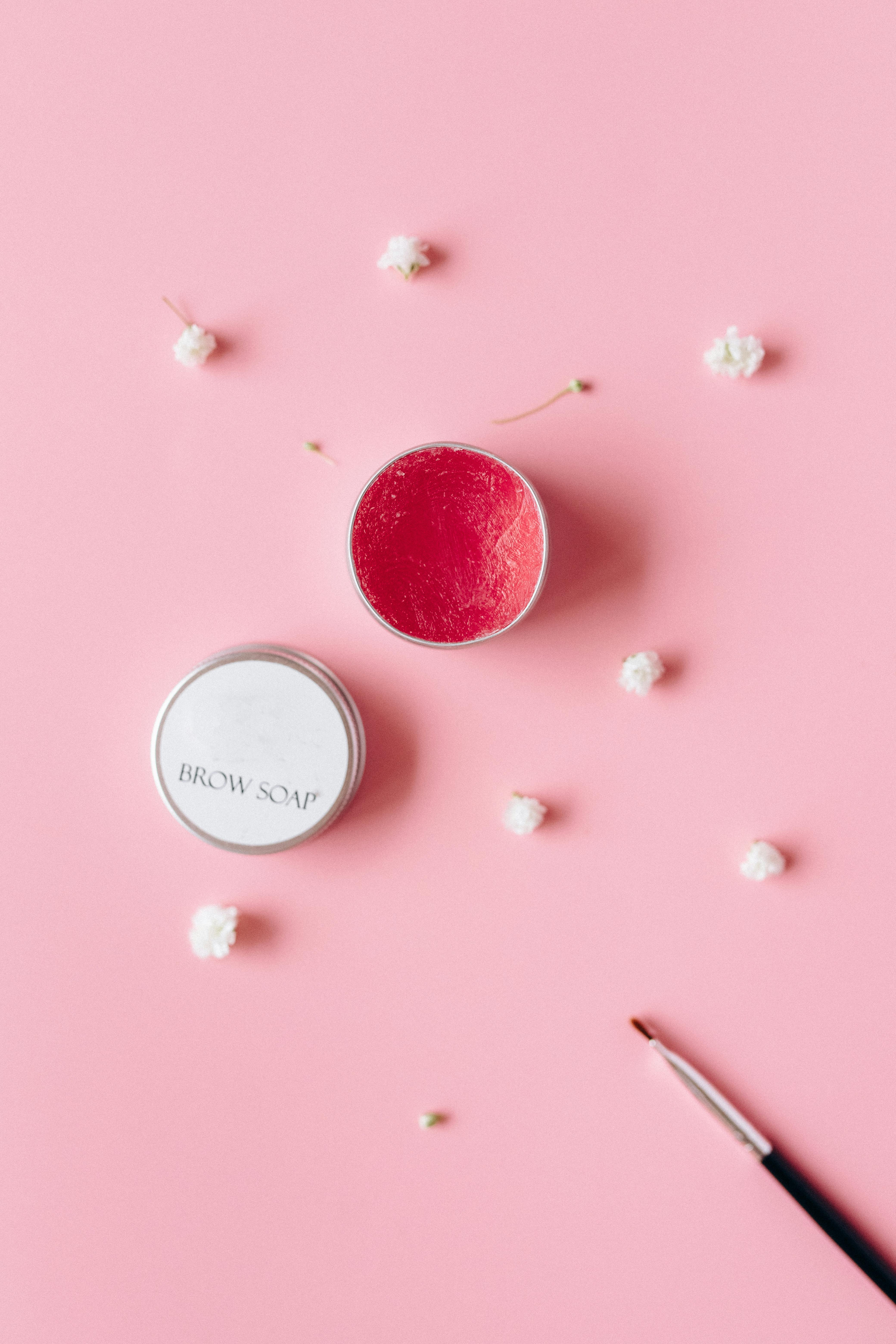Flat lay of brow soap and brush on a pink surface with small white flowers.