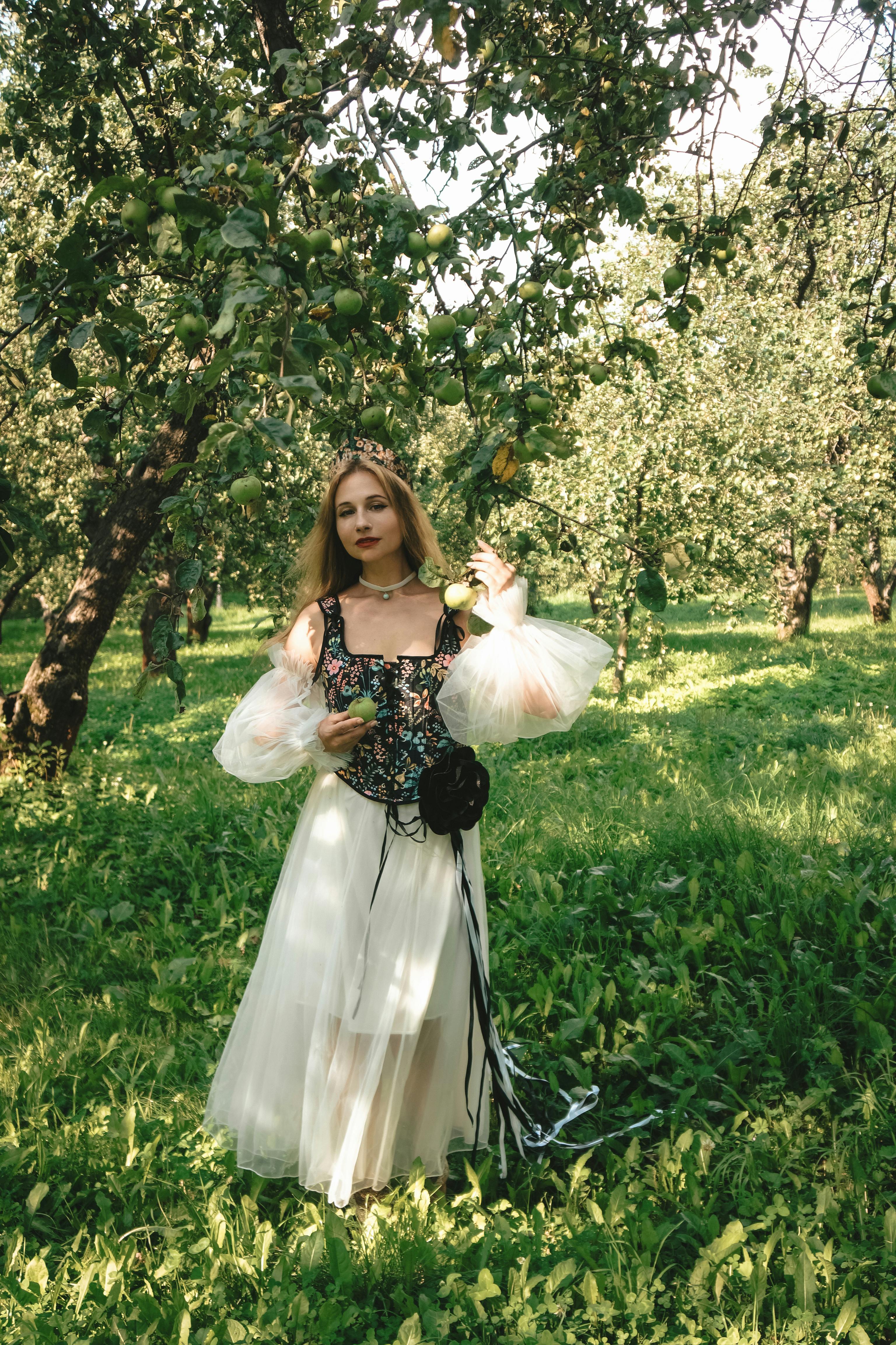 A woman in a white dress stands gracefully in a lush orchard during the day.