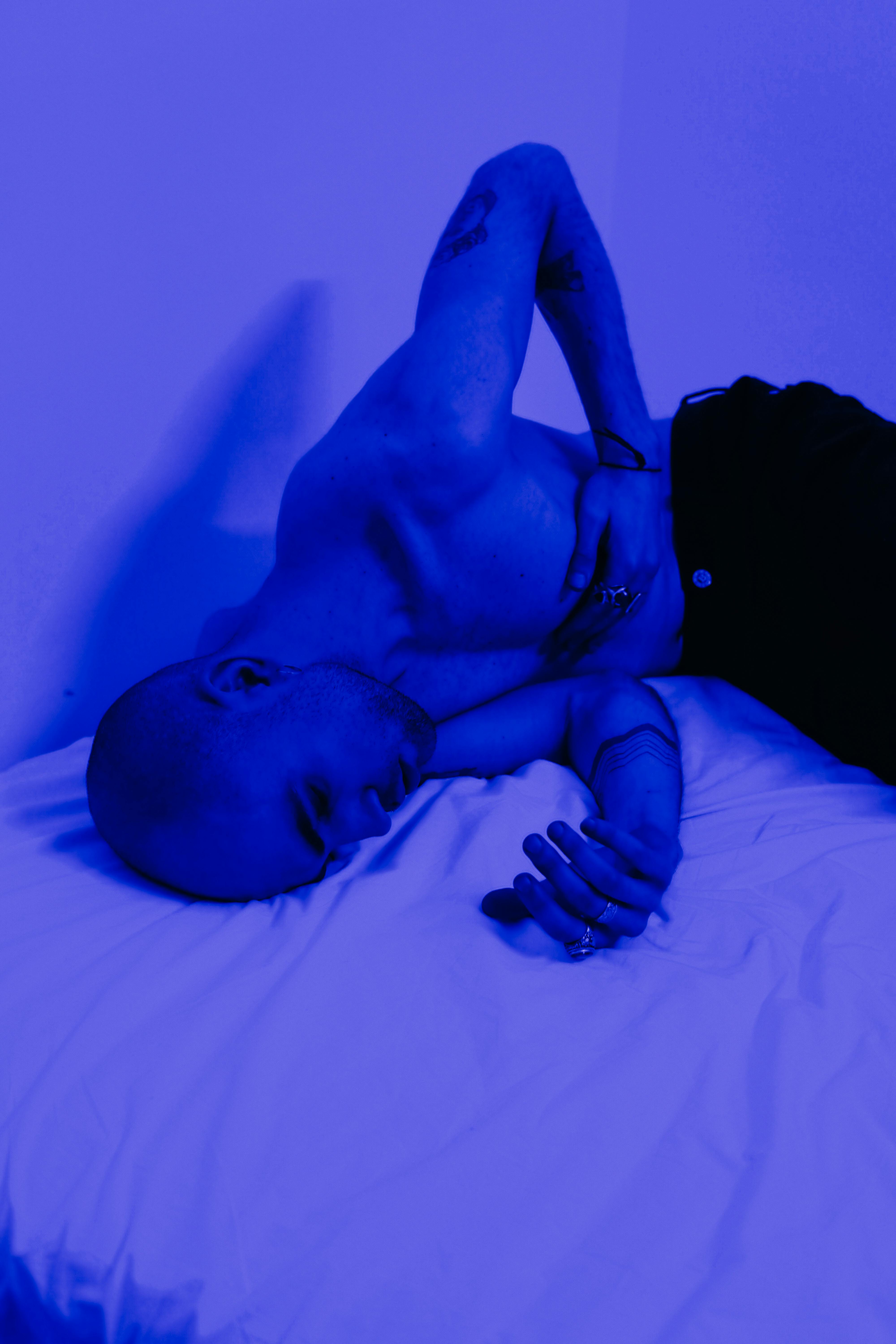 A shirtless man lies on a bed in blue light, expressing deep emotion and contemplation.