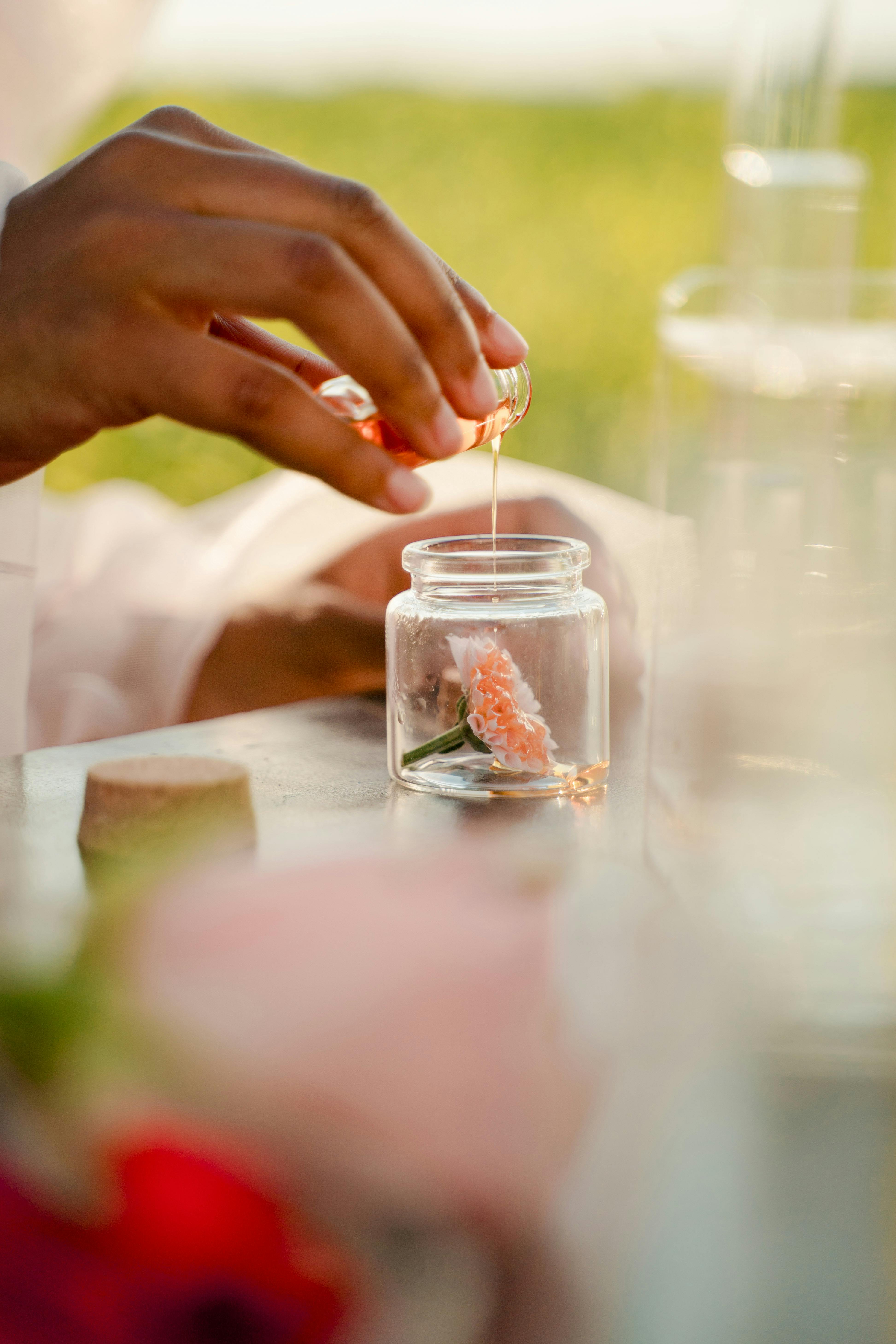 Close-up of hands pouring aromatic oil into a jar with a pink flower outdoors.