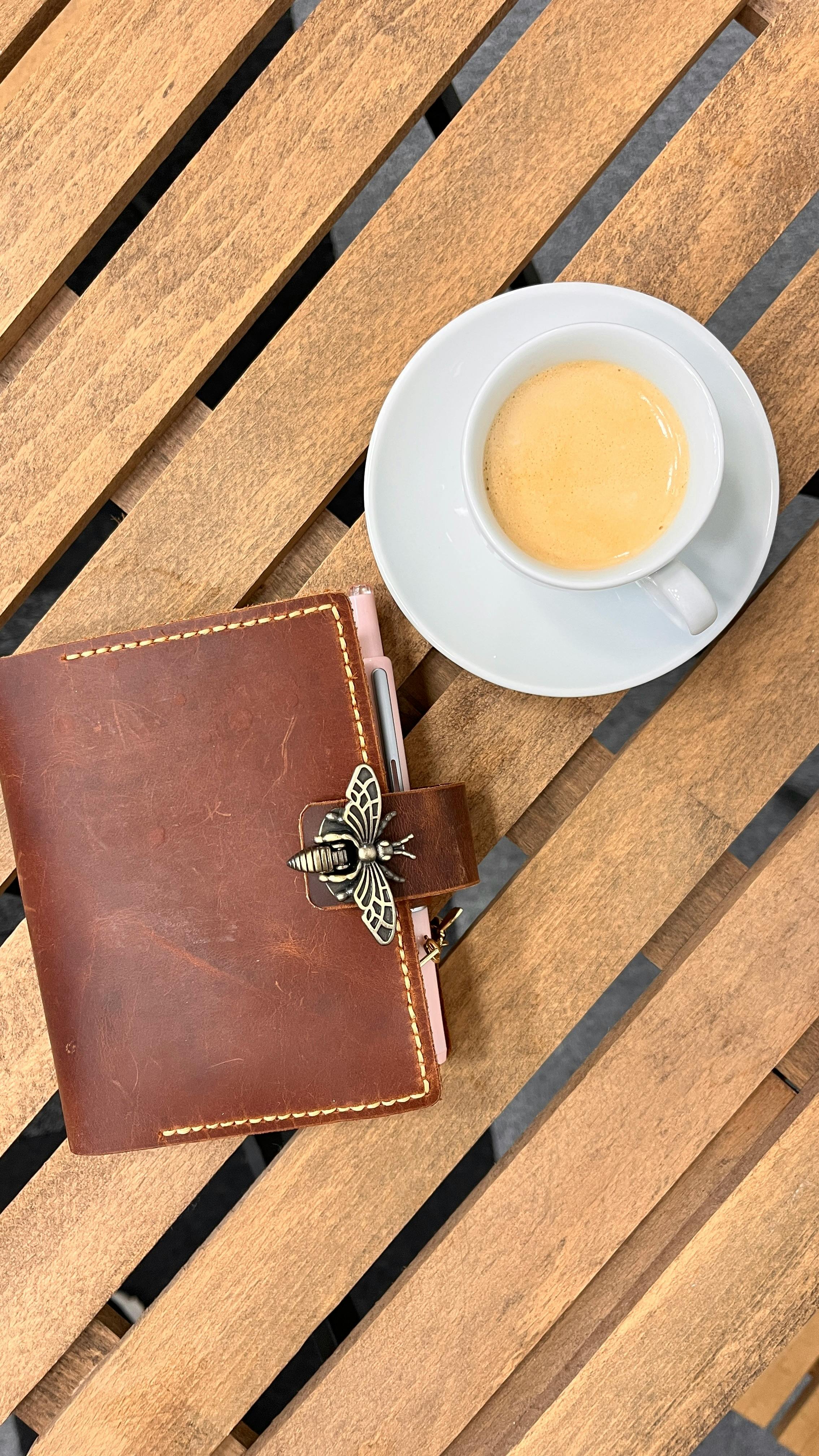 Top view of espresso and leather journal with bee logo on a wooden table.