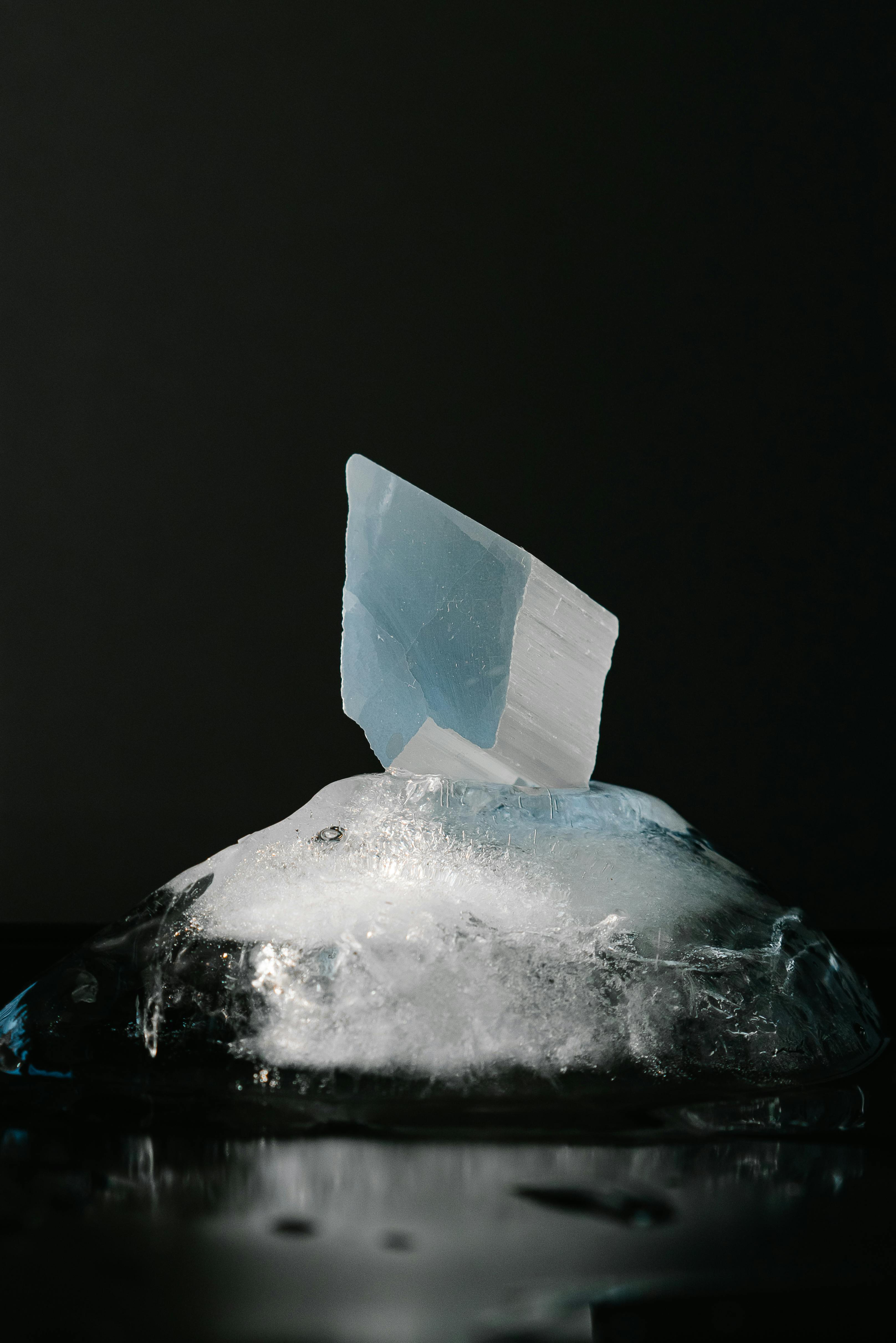 A shard of blue crystal emerging from a melting block of ice against a dark backdrop.