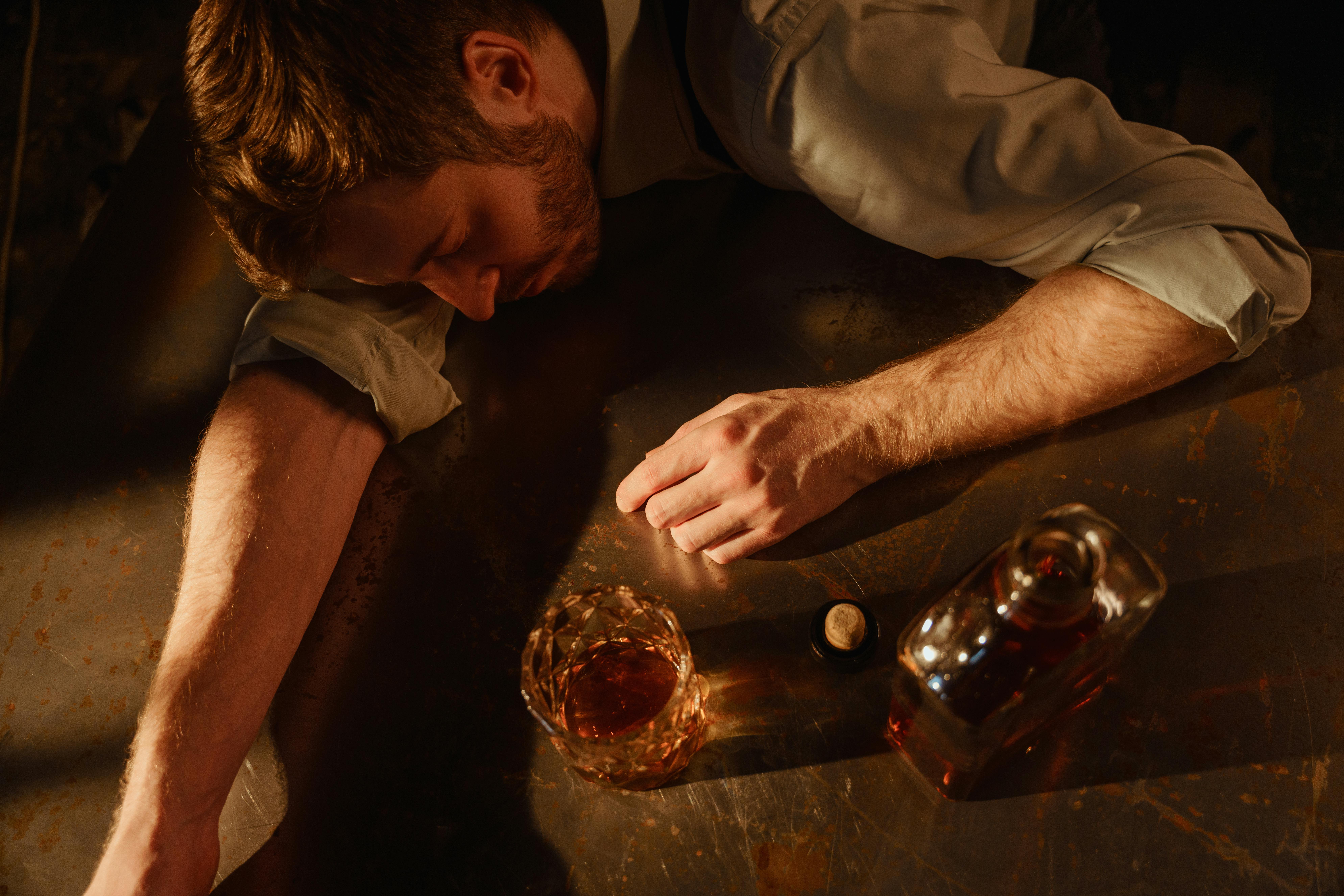 A man sleeps on a table with a whiskey glass and bottle nearby, suggesting exhaustion.
