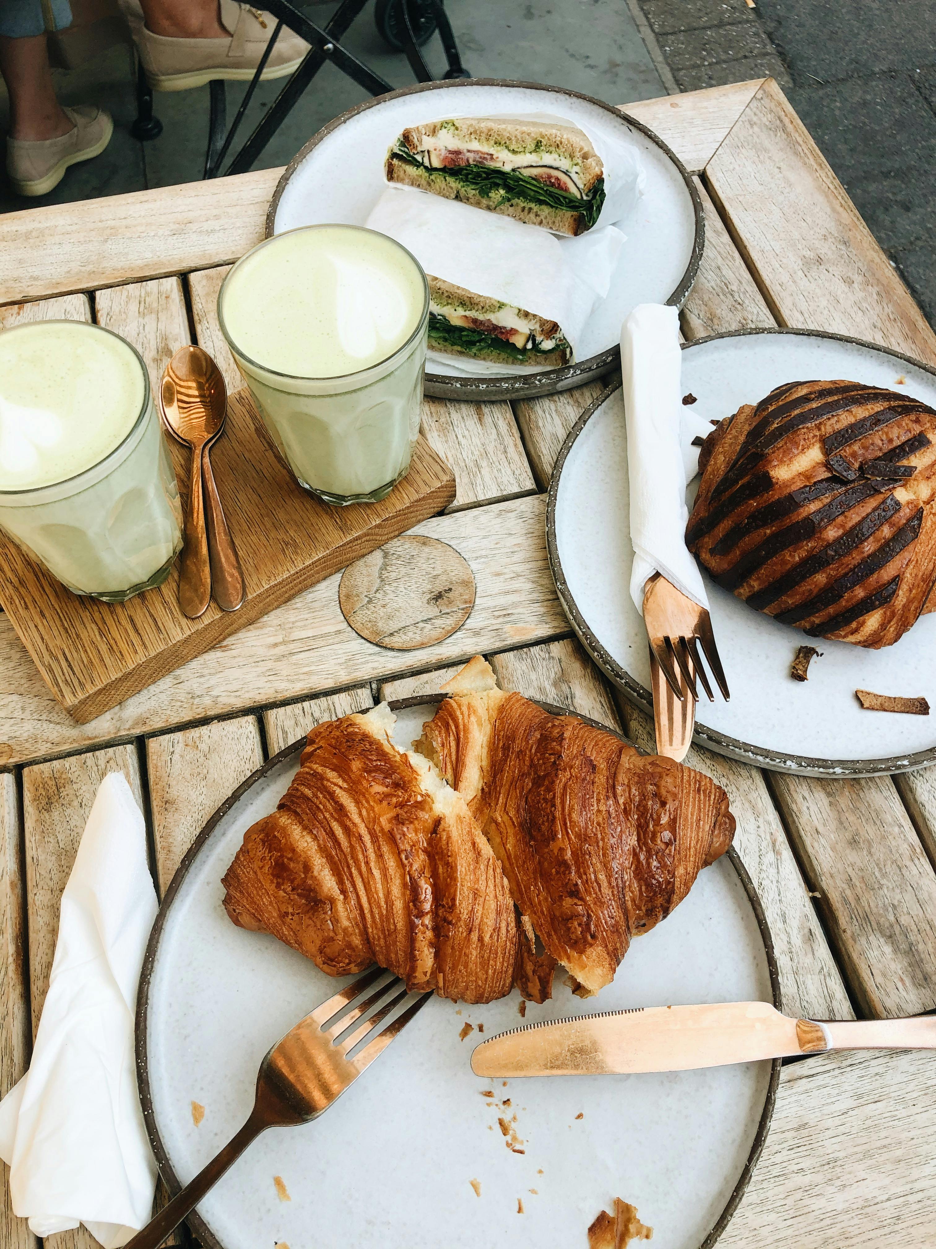 Experience a sophisticated brunch in London with croissants, sandwiches, and matcha lattes on a rustic wooden table.