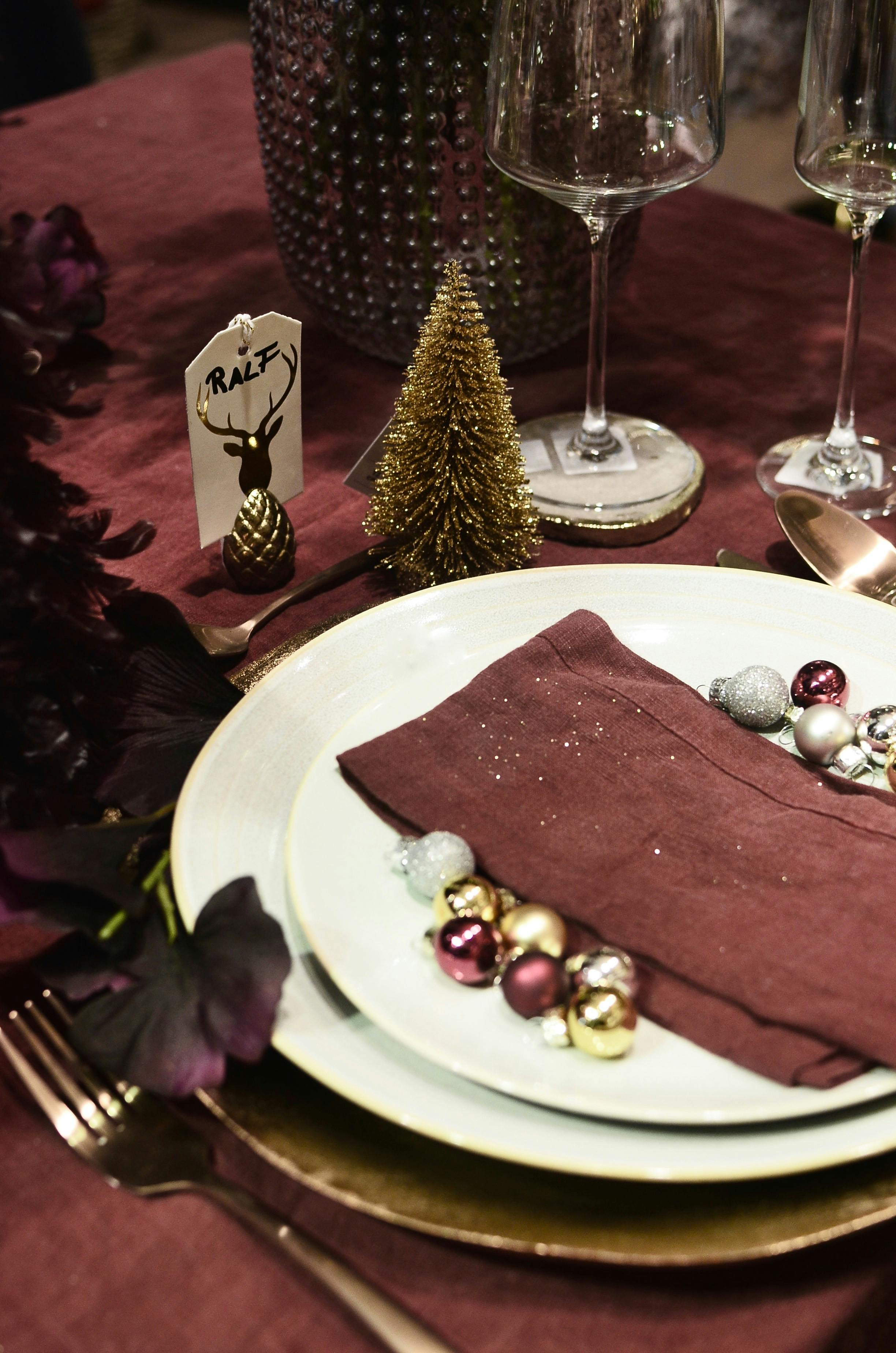 Festive holiday table setting with decorative baubles, napkins, and a miniature Christmas tree on a burgundy fabric.