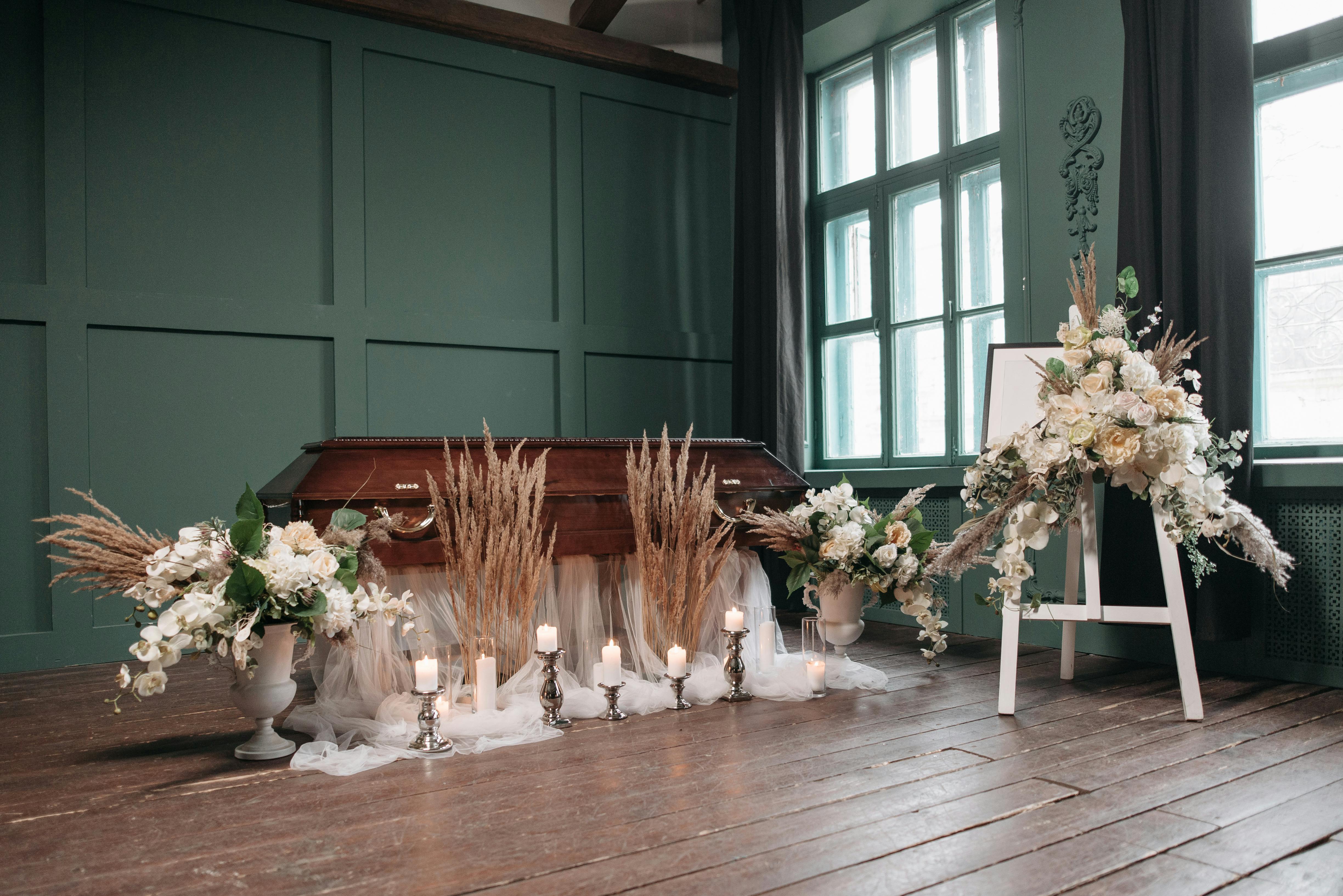 An indoor funeral setting with a wooden coffin, white flowers, and lit candles.