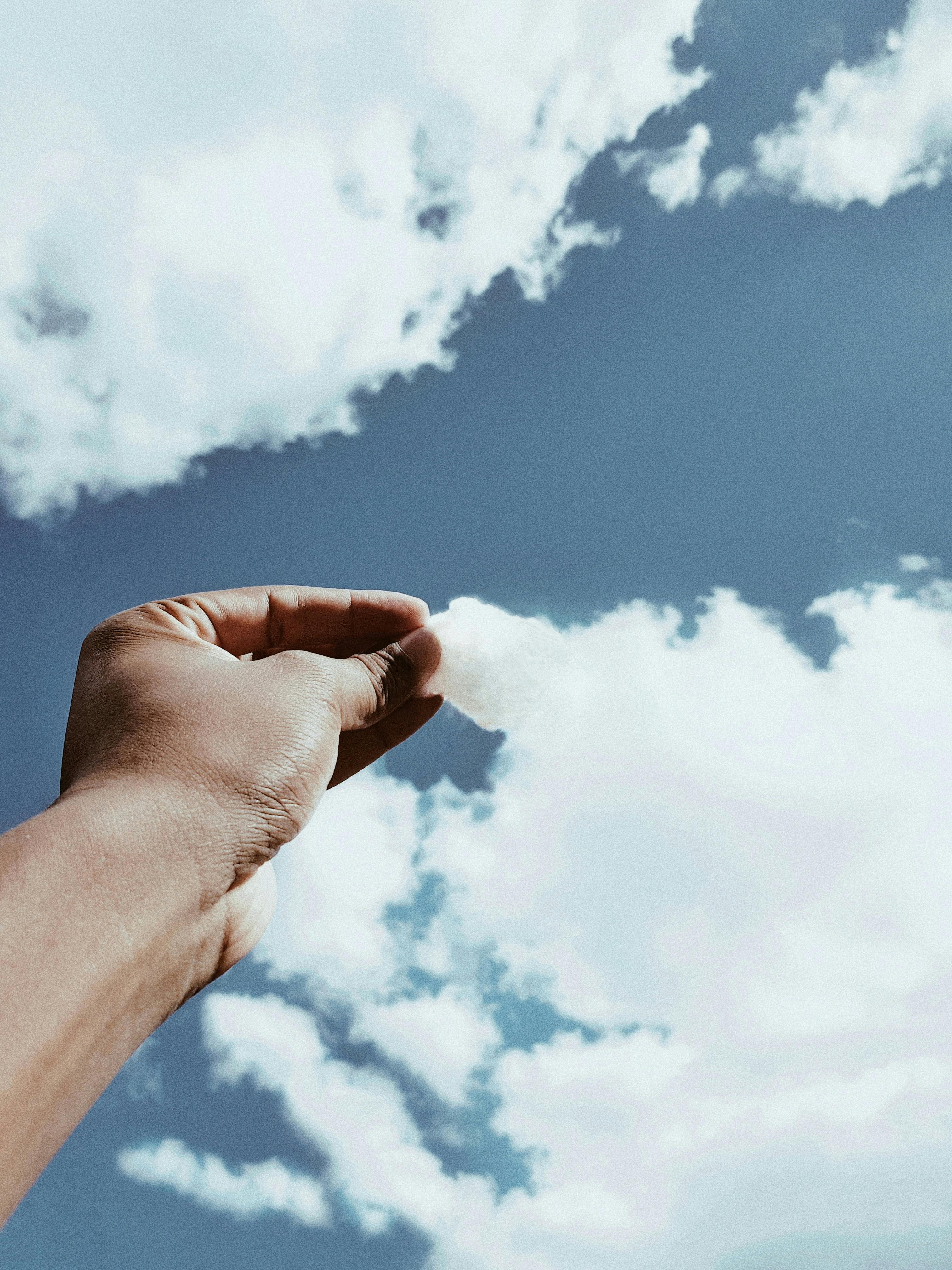 A hand trying to touch a cotton-like cloud against a bright blue sky, symbolizing dreams and aspirations.