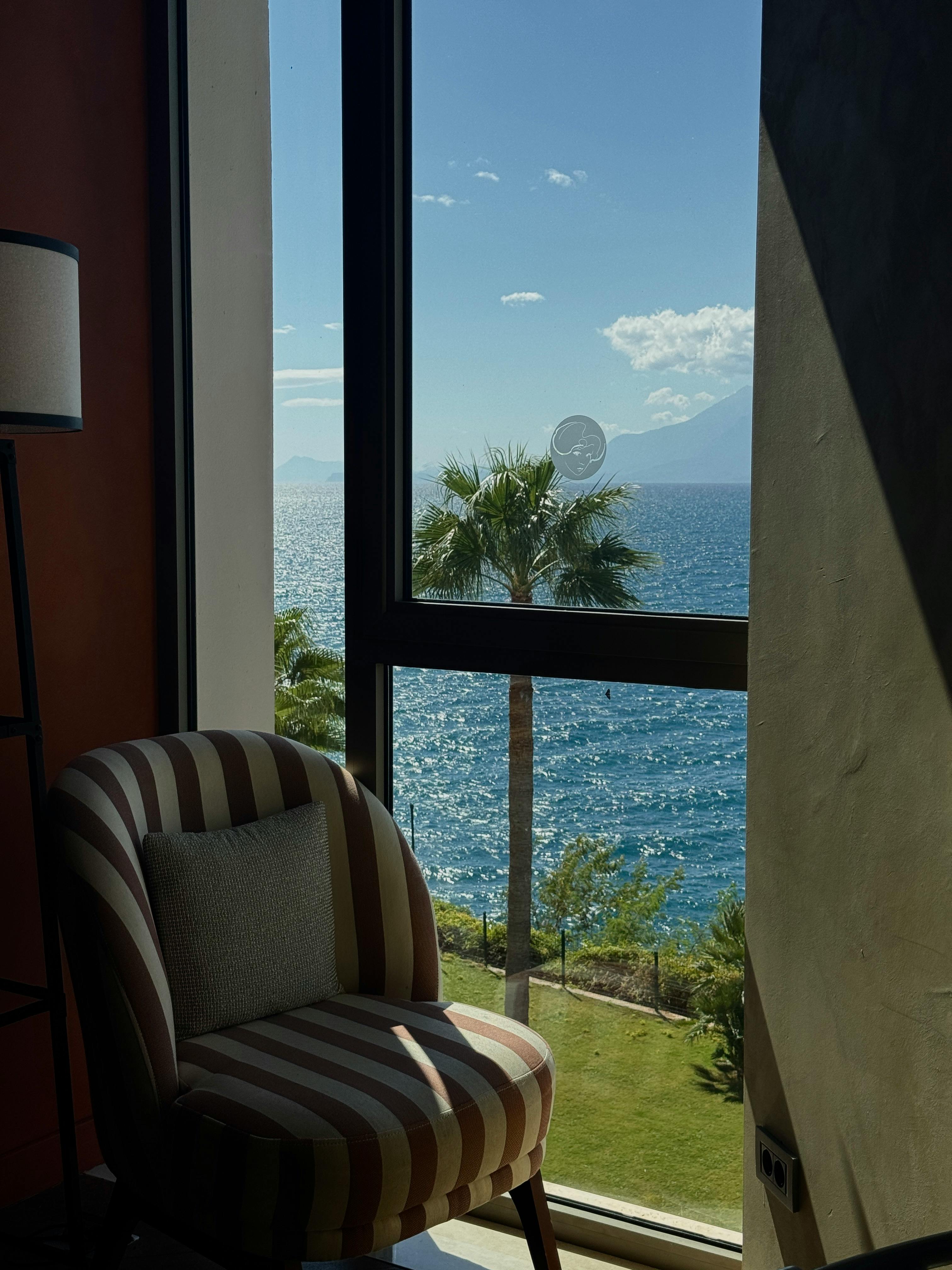 Sunlit room with striped chair overlooking ocean and palm trees.
