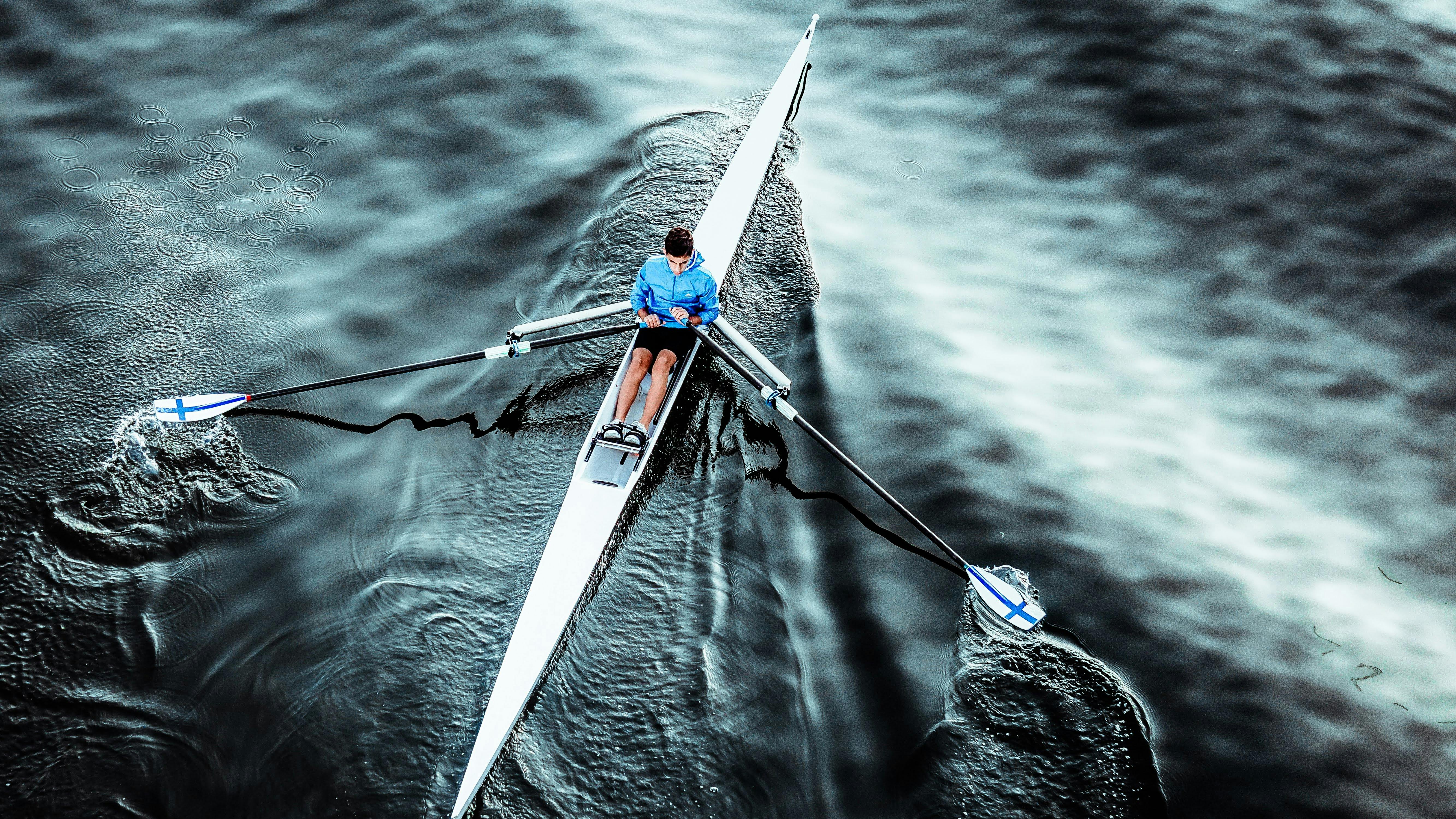 Aerial view of a man rowing a single scull on calm waters, dressed in a blue jacket.