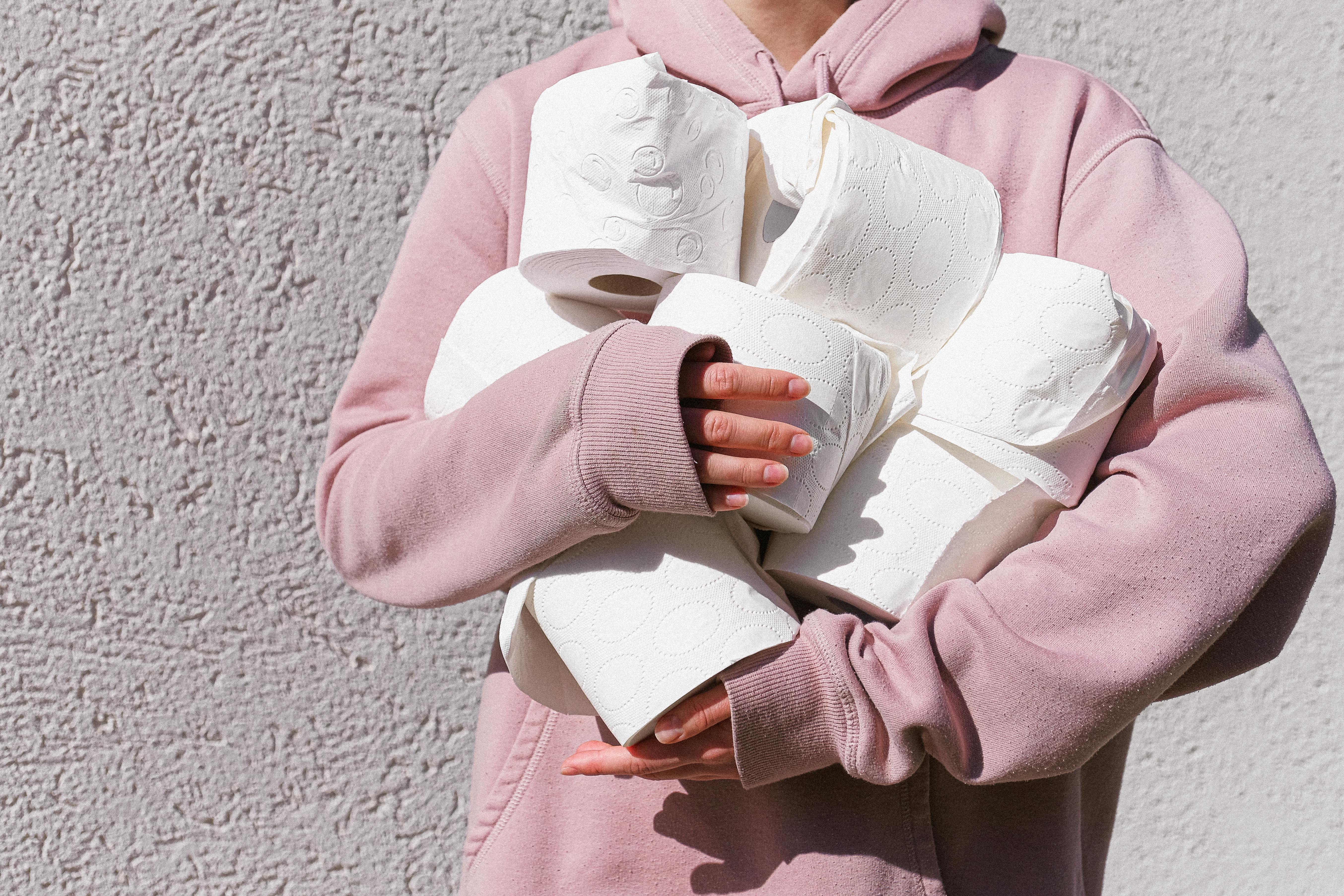 A person in a pink hoodie holds multiple rolls of toilet paper against a textured wall.