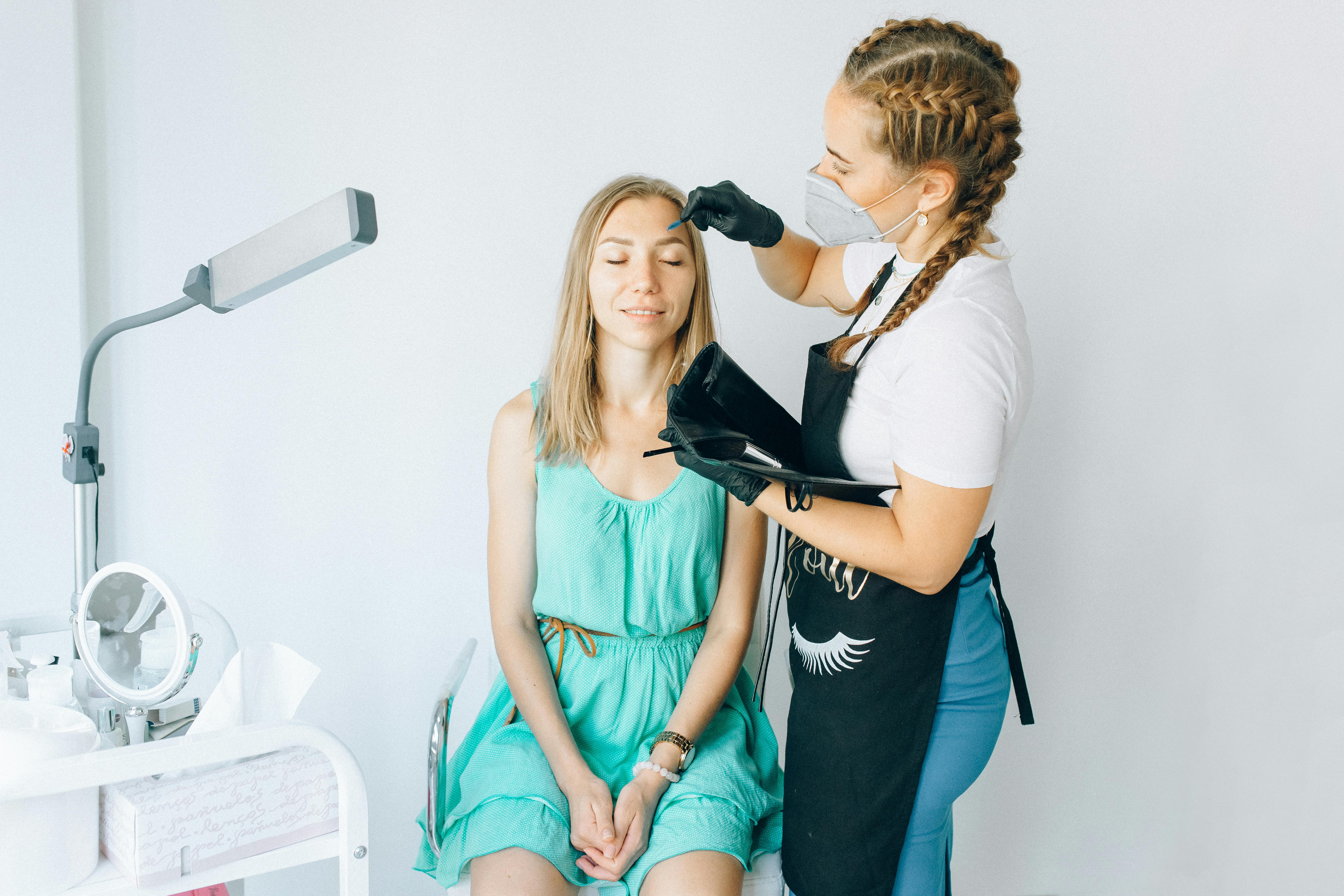 A beautician performs eyebrow microblading on a woman in a modern salon setting.