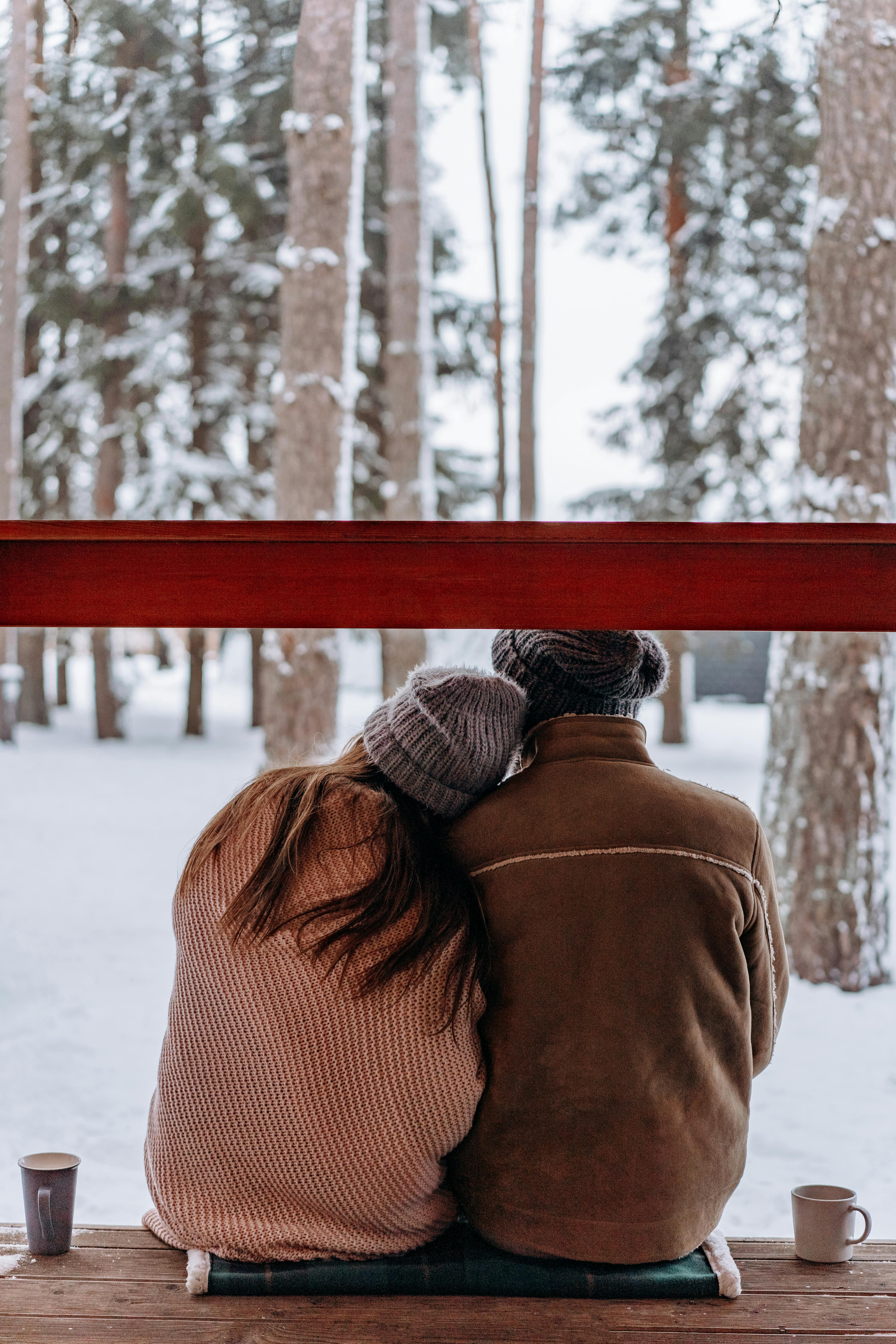 A couple in cozy winter attire sitting together in a snowy forest, enjoying a warm drink.