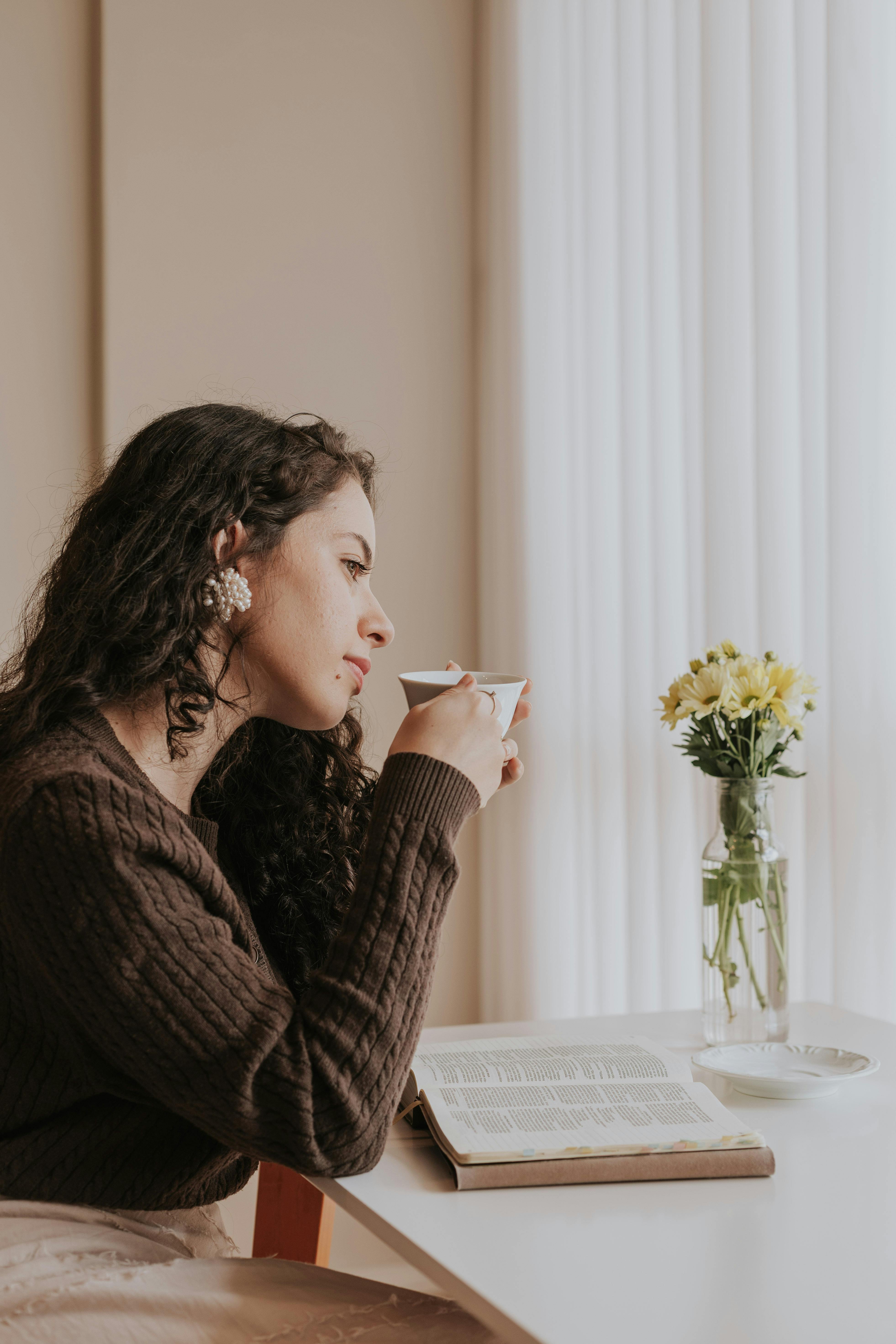 A woman enjoys a quiet moment sipping coffee at home, with a book and flowers on the table.