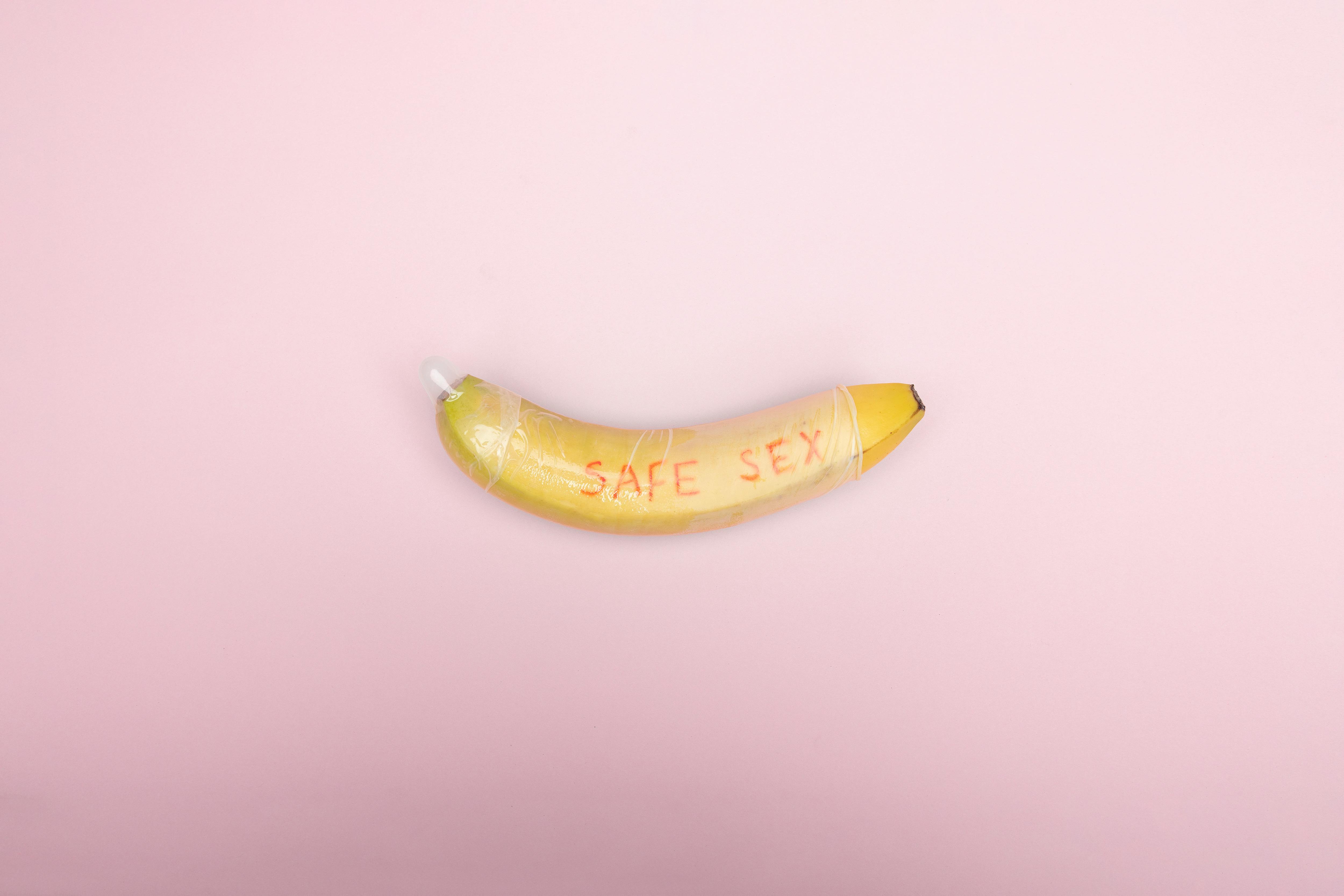Still life image of a banana wrapped in a condom with 'safe sex' text on a pink background.