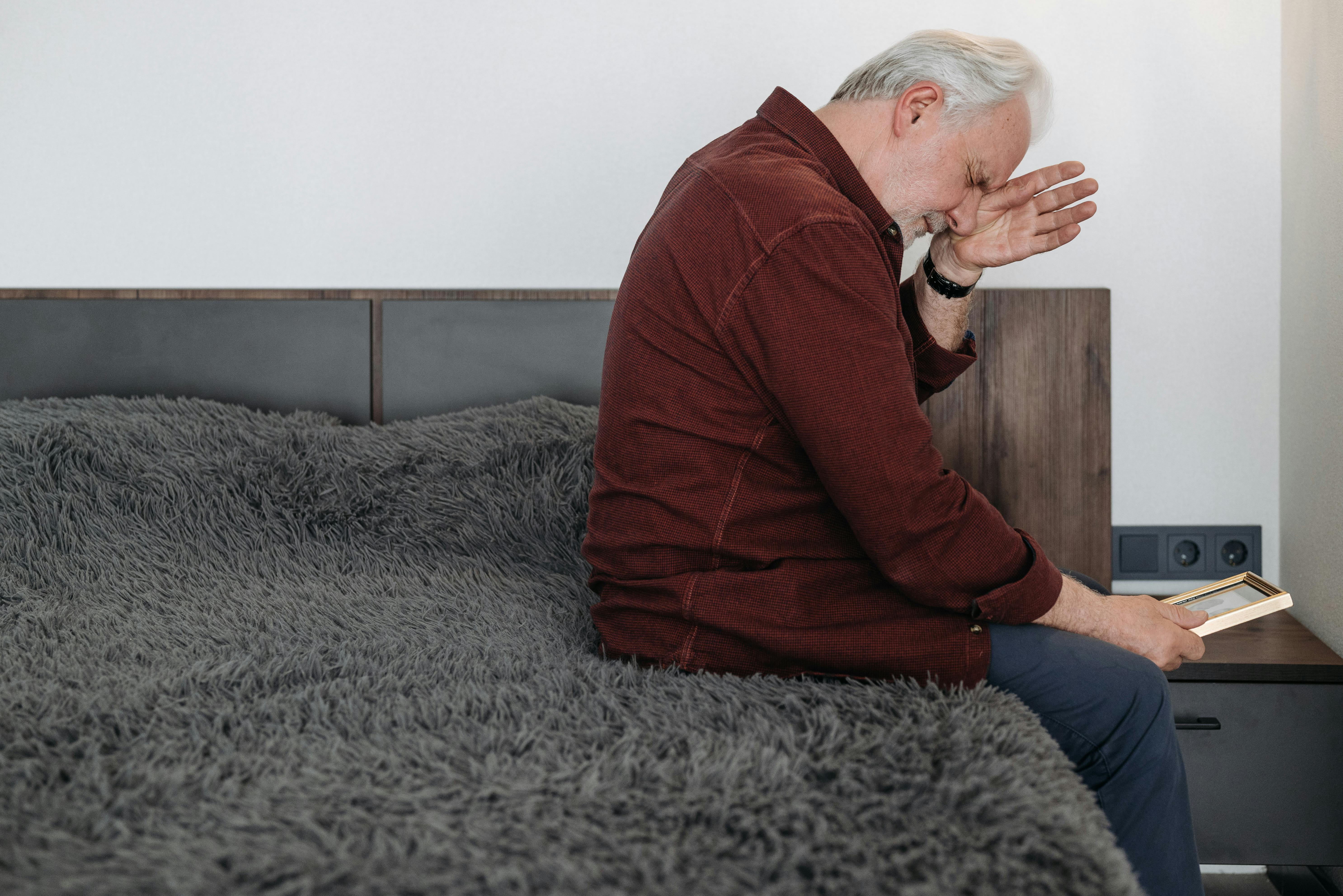 An elderly man sits on a bed in tears holding a picture frame, depicting bereavement.